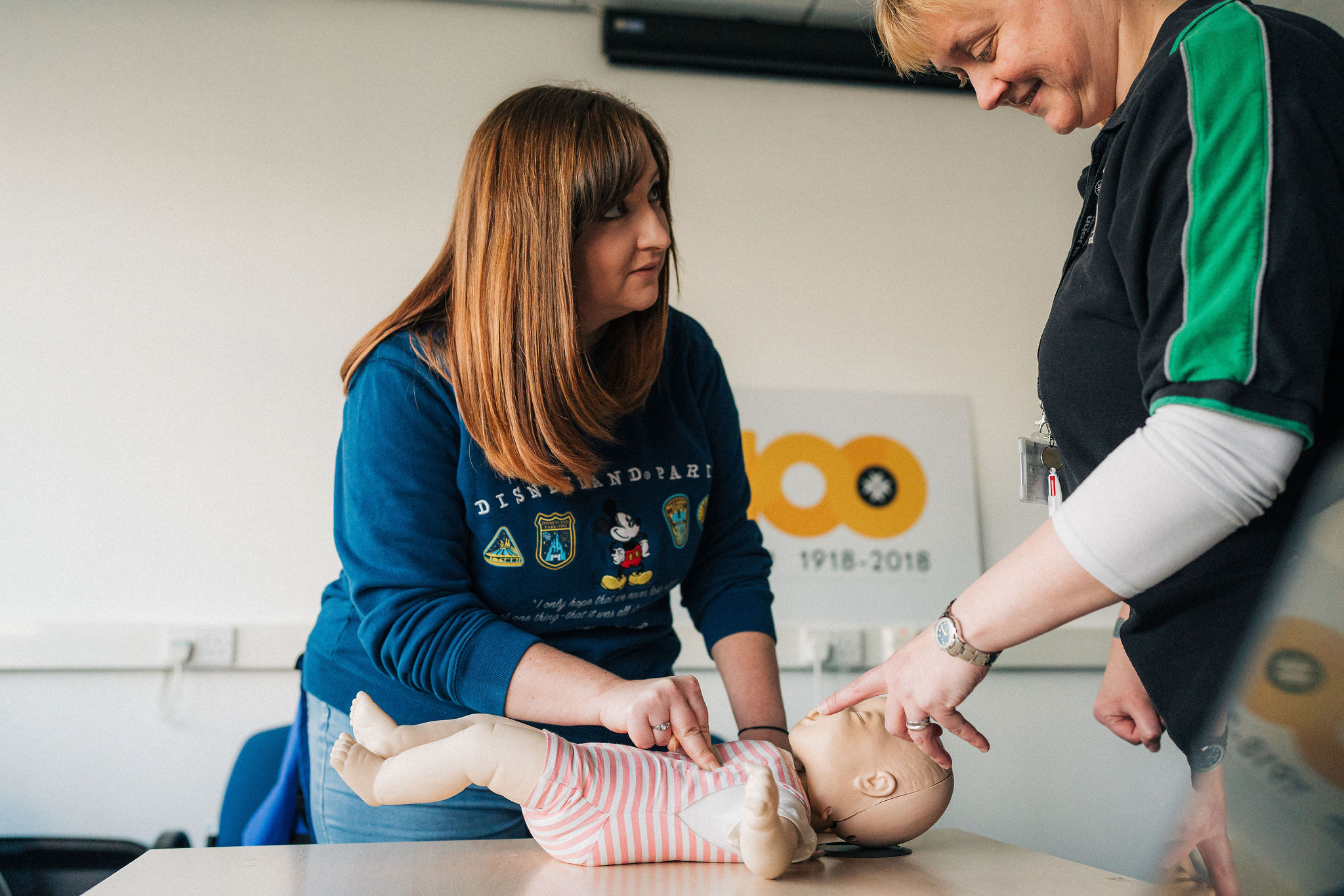 A St John Ambulance Cymru course trainer is teaching a delegate how to do CPR on an infant manikin. The delegate has placed their index and middle finger in the centre of the infant manikin's chest.
