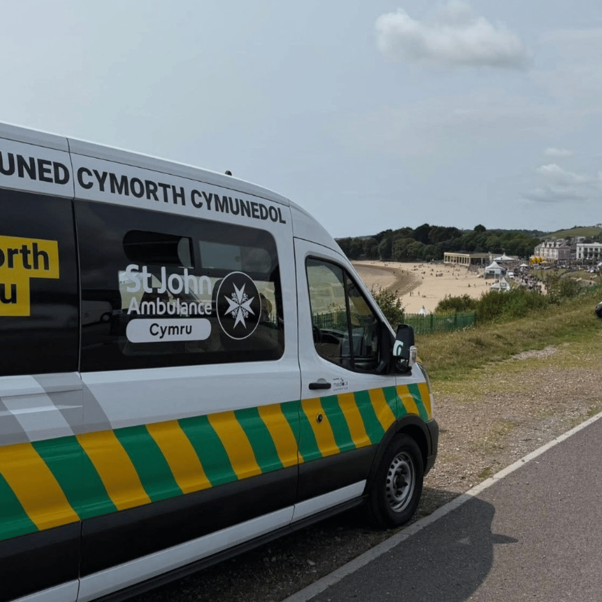 The side view of an ambulance parked alongside Barry beach. The side window has the St John Ambulance Cymru logo printed over it. The vehicle is white with a strip of green and yellow diagonal stripes running the length of the vehicle.
