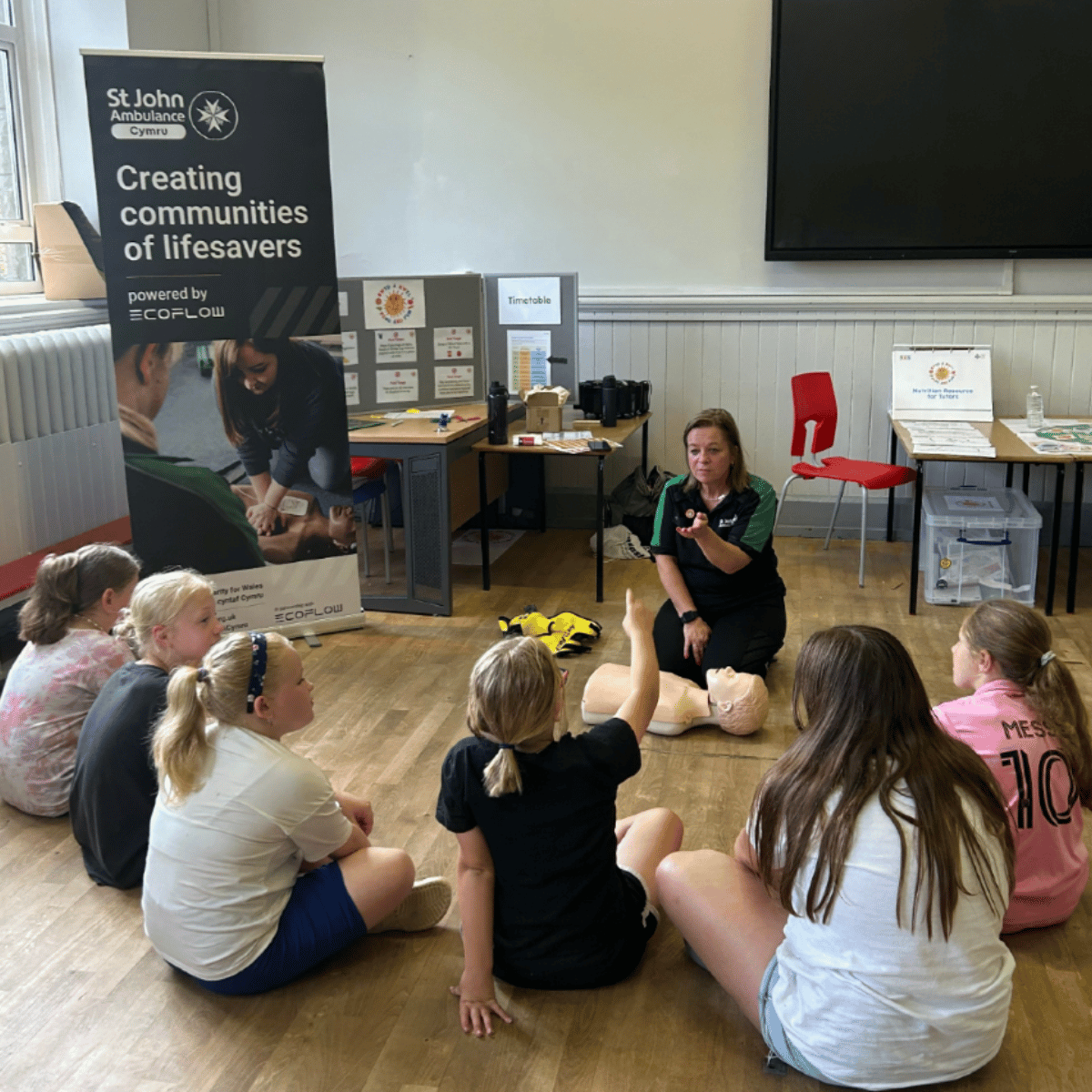 A St John Ambulance Cymru volunteer is kneeling on the floor with a CPR training manikin laid out in front of her. Circled around her are little girls, who are sat cross legged and watching the first aid demonstration. They appear to be in a classroom setting.