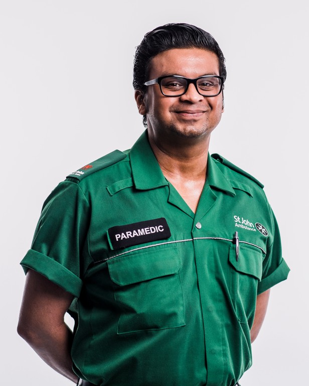 A St John Ambulance Cymru volunteer standing with his arms behind his back. He is wearing a green shirt with the St John Ambulance logo and a badge that reads 'Paramedic.' He is smiling.