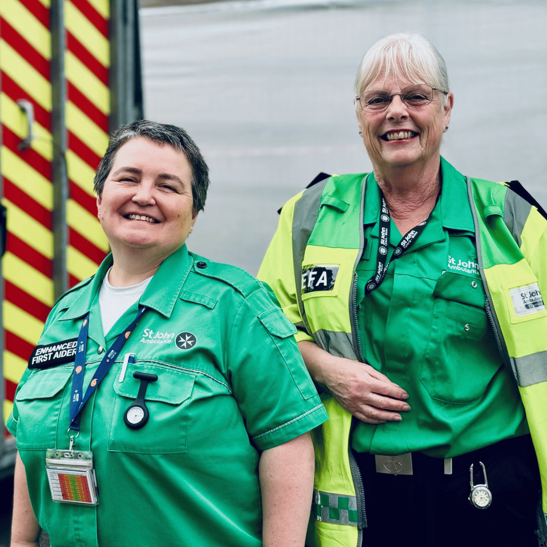 Two St John Ambulance Emergency First Aiders posing for a photo wearing their green St John Ambulance Cymru uniform. Both are smiling and an ambulance is in the background.