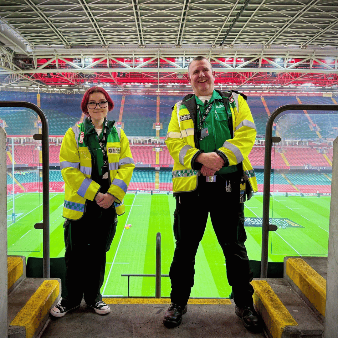 Two St John Ambulance staff are standing near the top seats of the Principality Stadium, with the Rugby pitch behind them. Both are wearing our green uniform and bright yellow high visibility jackets with the St John Ambulance Cymru logo on the front of their jackets.