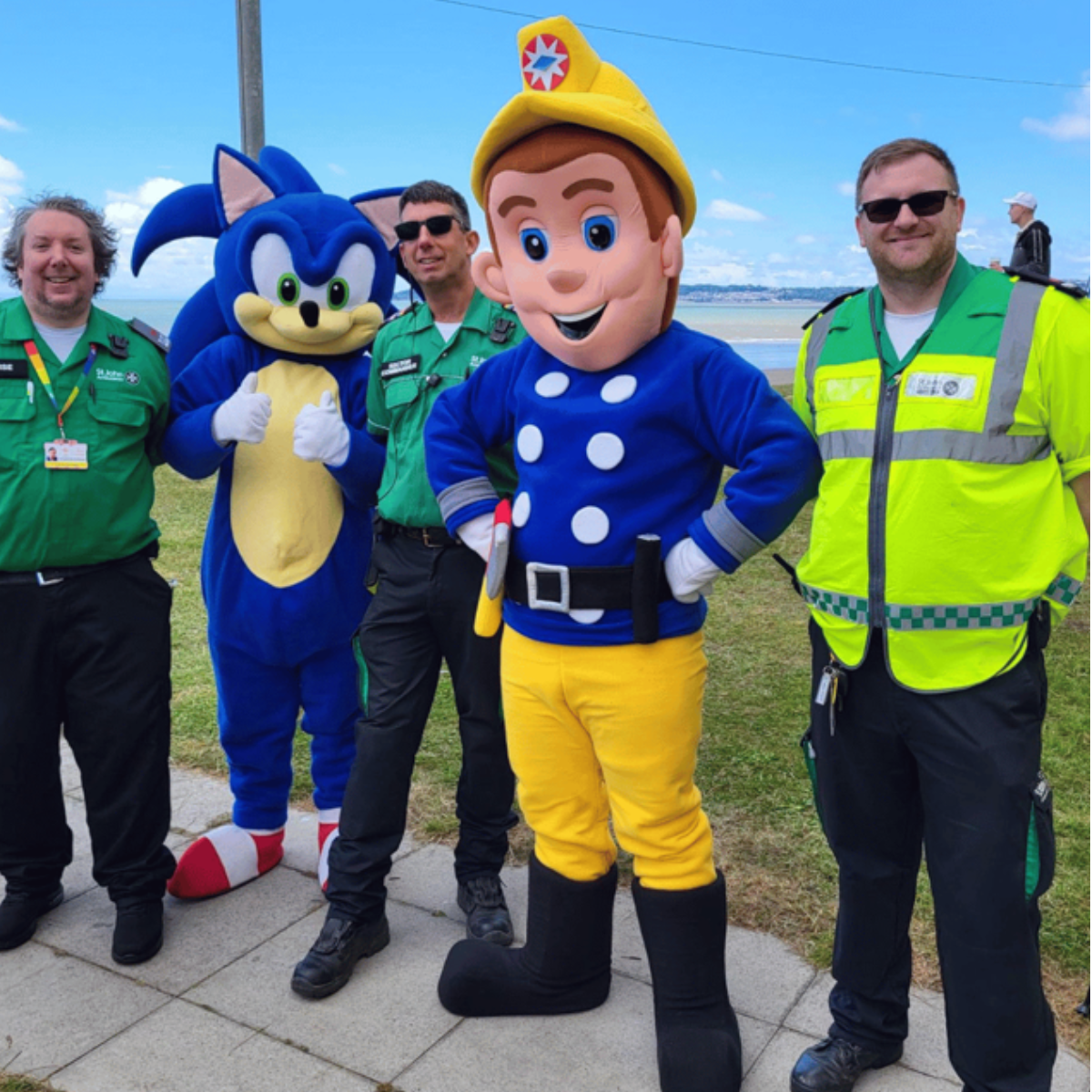 three men in paramedic uniforms pose outdoors on a paved area near grass and water, with two people in character costumes standing between them. One costume is of Sonic the Hedgehog, giving two thumbs up, and the other is Fireman Sam with hands on hips. The sky is blue with some clouds, and there is a coastline in the background.