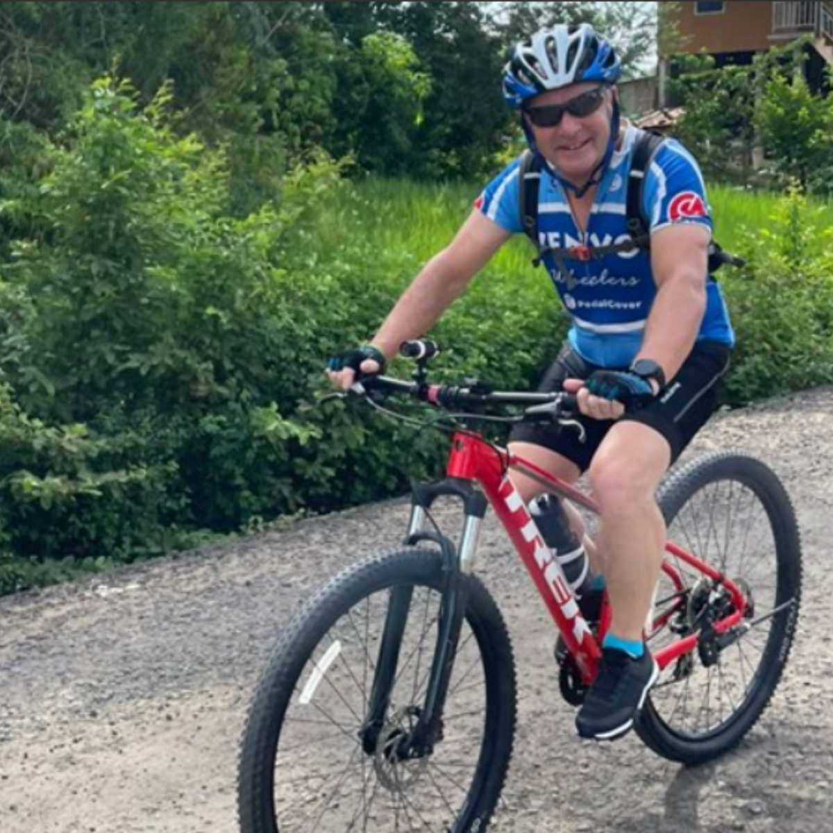 Colin wearing a blue cycling jersey, black shorts, a helmet, and sunglasses rides a red mountain bike on a gravel path. He is smiling and appears to be enjoying the ride. He is outside.