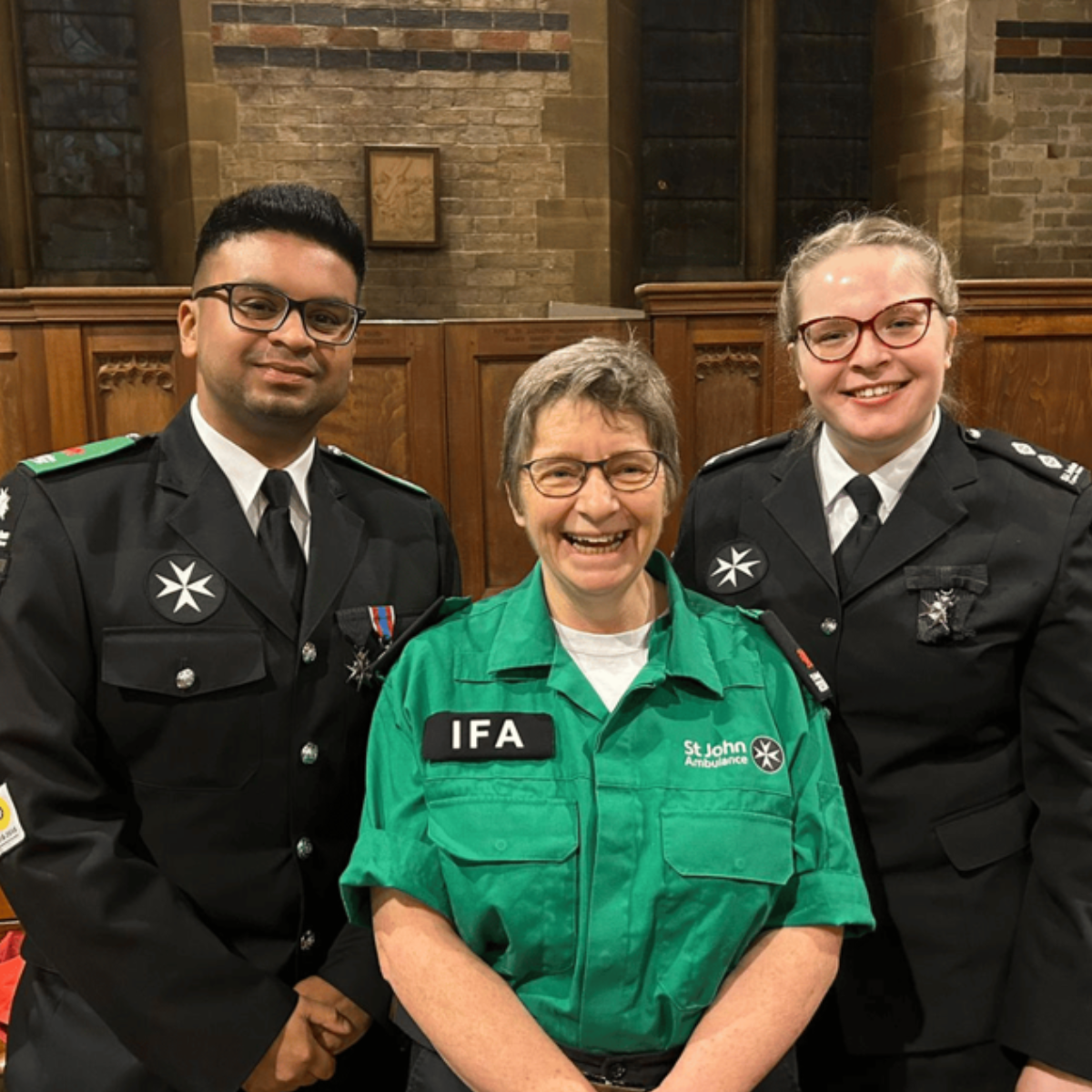 Three St John Ambulance Cymru volunteers standing next to each other and smiling. They appear to be in a church setting. All are wearing uniform and two of them have medals on their front pockets.
