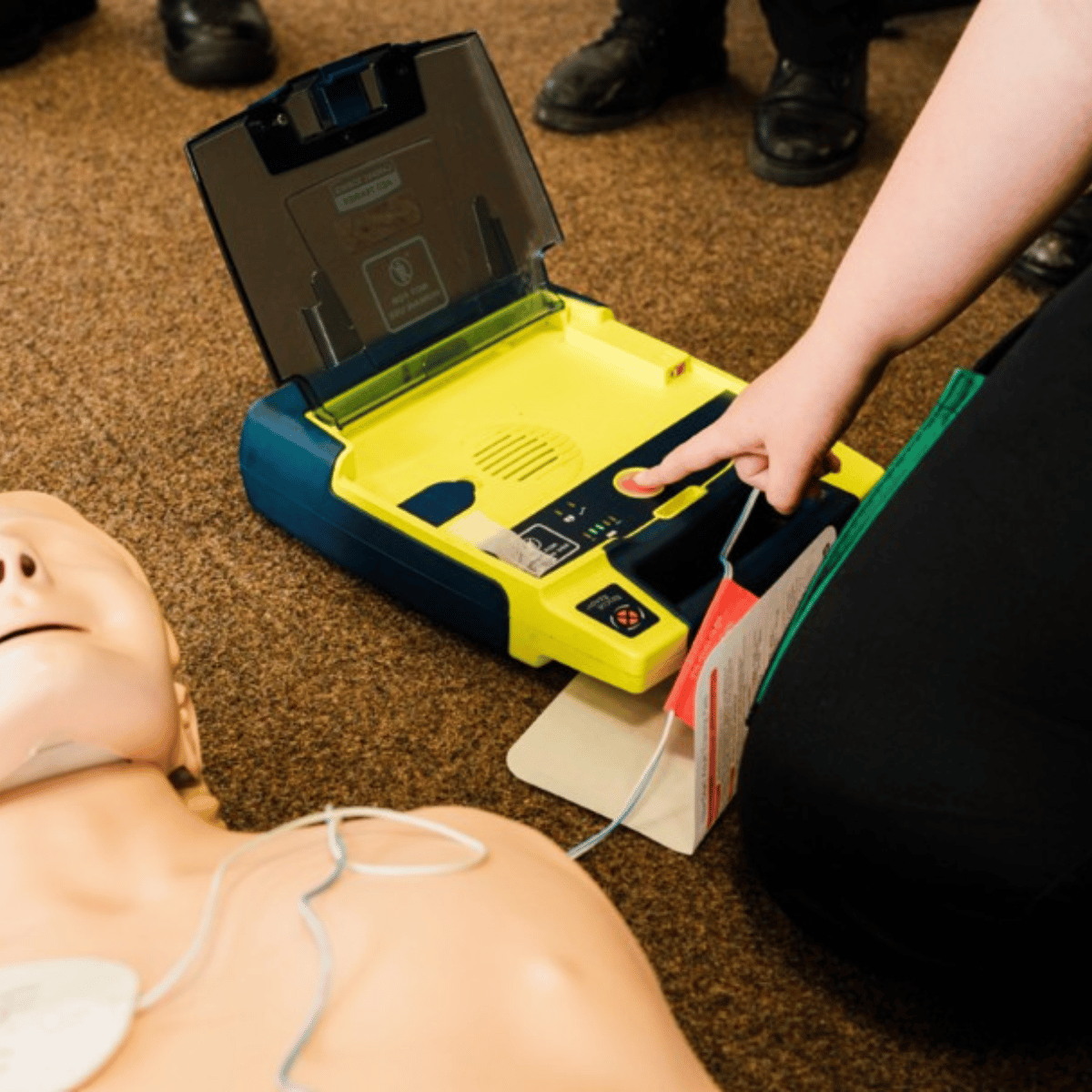 A yellow defibrillator indoors attached to a manikin with pads, with a person ready to deliver a shock.