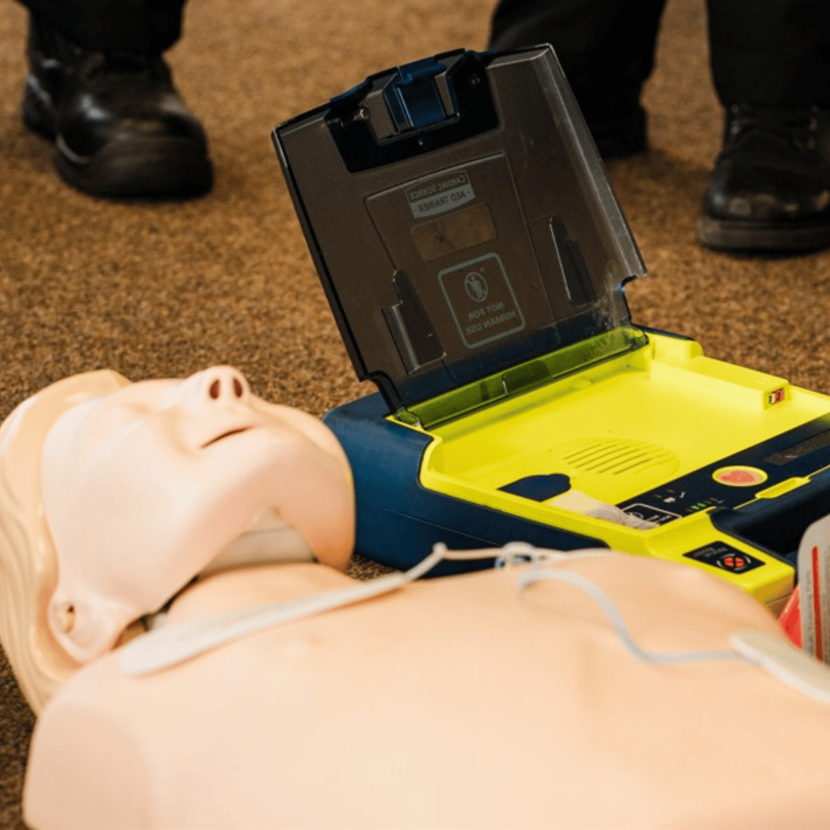 A yellow defibrillator indoors attached to a training manikin with pads.