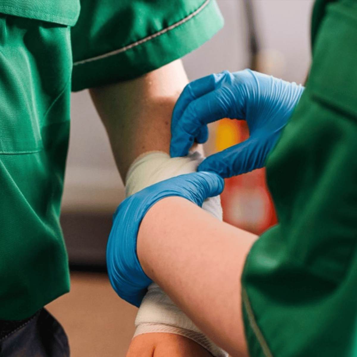 Two St John Ambulance Cymru staff in green uniforms. One is wearing blue gloves and is applying a bandage to the arm of the other.