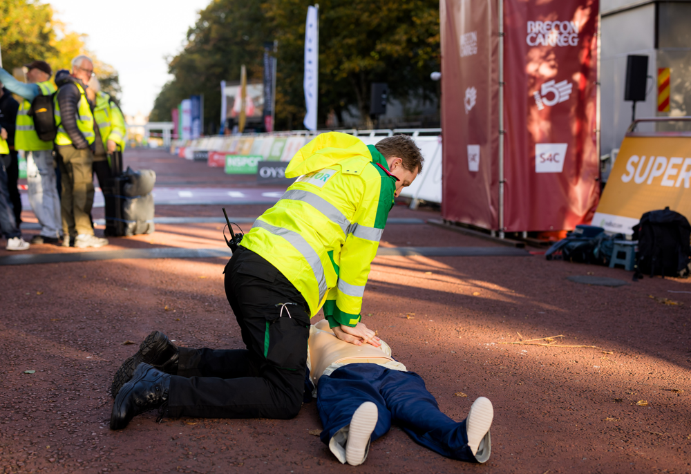 Volunteer Doctor in a high visibility St John Ambulance Cymru coat giving CPR to a training manikin at the Cardiff Half marathon finishing line.