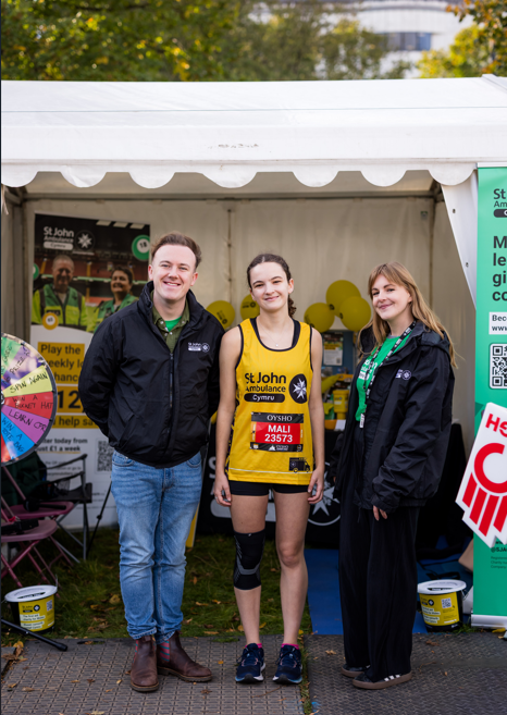 Two St John Ambulance Cymru staff members standing with a runner from the Cardiff Half Marathon. The runner is wearing the St John Ambulance Cymru vest, which fundraisers for St John Ambulance Cymru wear for the race. They are all smiling.