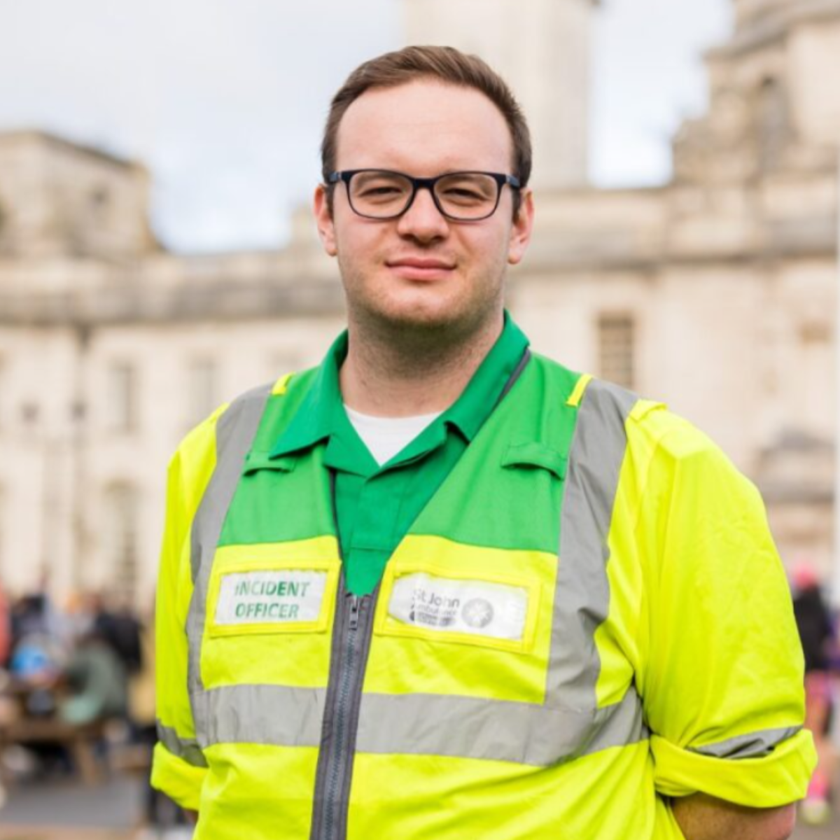 A volunteer smiling. They are wearing a green high visibility coat. On the chest area of the coat, there is a St John Ambulance Cymru logo and a badge with the wording 'incident officer'. They are standing outside.