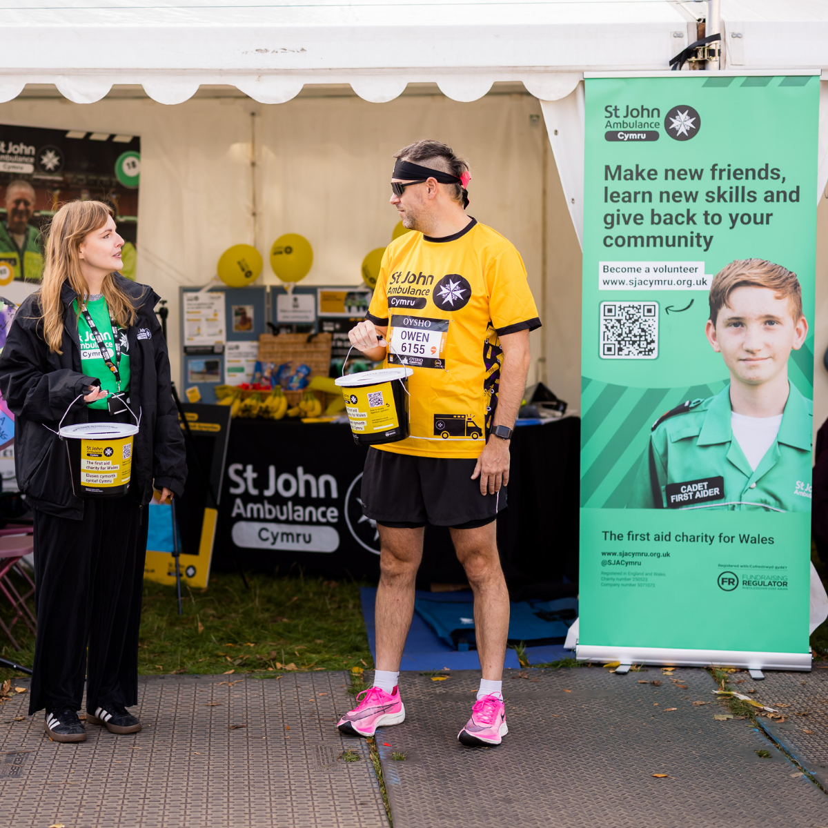St John Ambulance Cymru staff members holding out donation buckets. They are standing in front of an event tent and behind them is a table with St John Ambulance Cymru merch on and a table cloth with our logo.