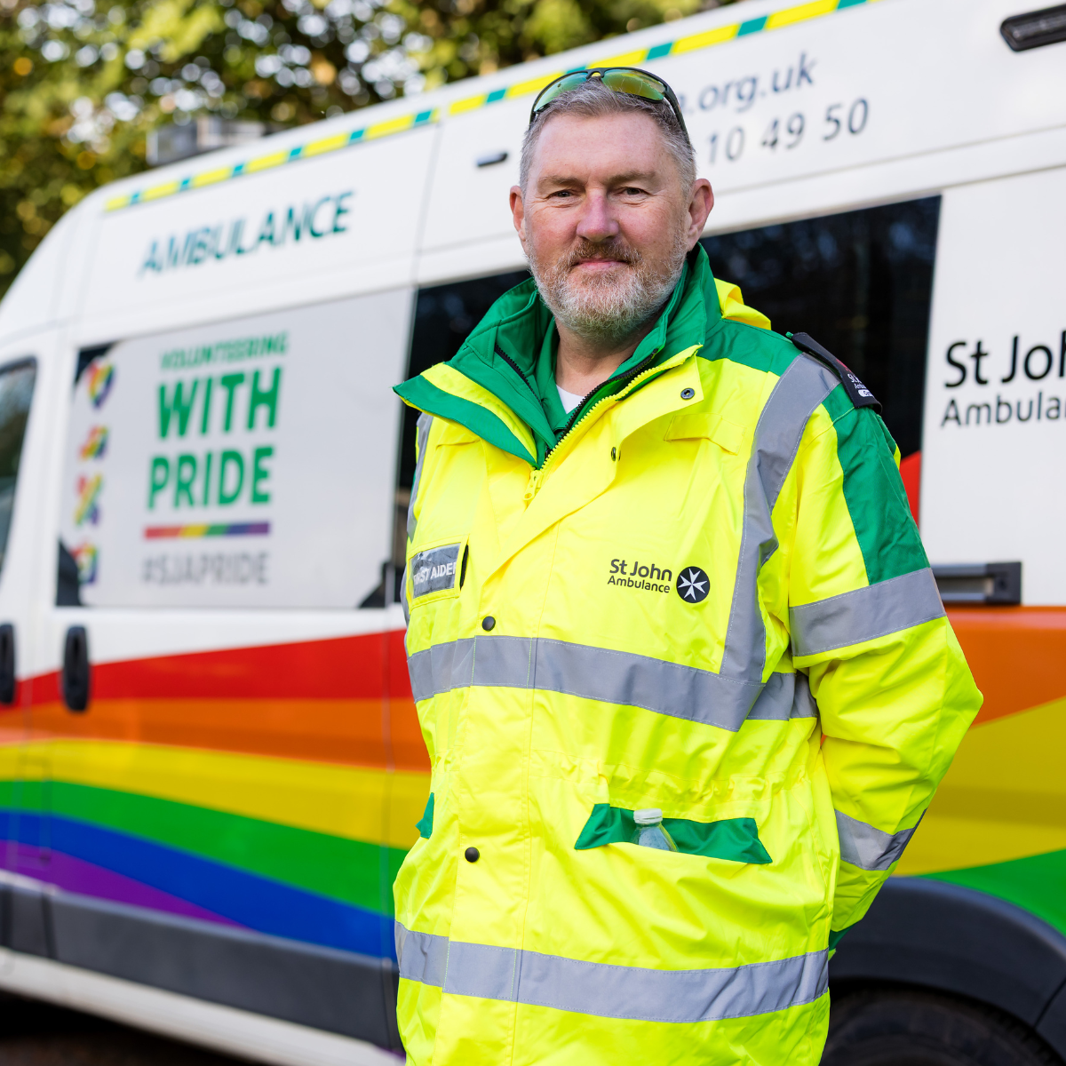 A volunteer smiling. They are wearing a green high visibility coat with the St John Ambulance Cymru logo and first aider badge. They are standing in front of one of our ambulances.