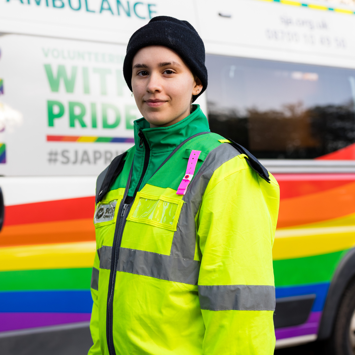 A volunteer smiling. They are wearing a green high visibility coat with the St John Ambulance Cymru logo and a black hat. They are standing in front of one of our ambulances.