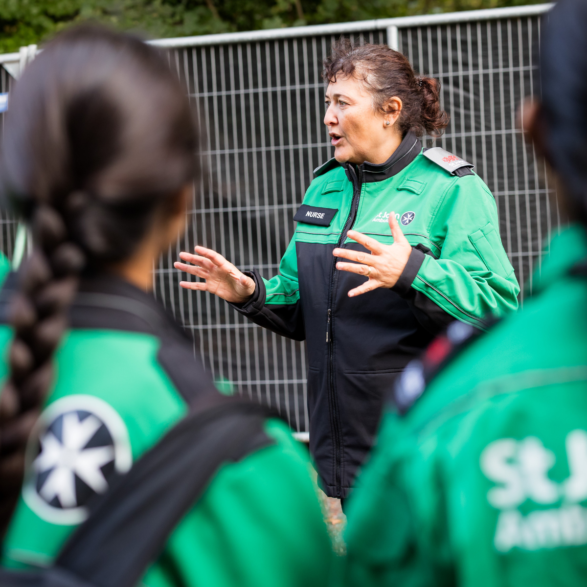 A volunteer nurse speaking to other St John Ambulance Cymru volunteers. She is wearing green and black uniform and is in mid conversation, gesturing with her hands. She has a badge with the wording Nurse on her coat.