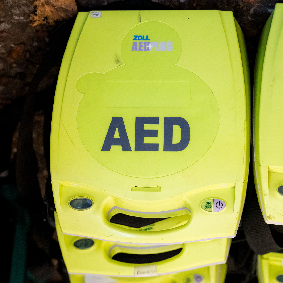 A bright yellow AED (Automated external defibrillator). There are 3 stacked on top of each other and are on the ground outside.