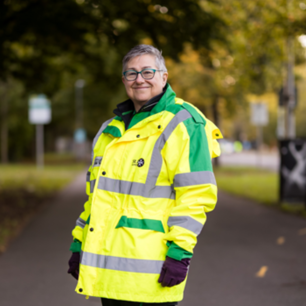St John Ambulance Cymru volunteer smiling for the camera. She has grey hair and blue glasses. She is wearing a high visibility bright green coat with the St John Ambulance logo printed on the front.