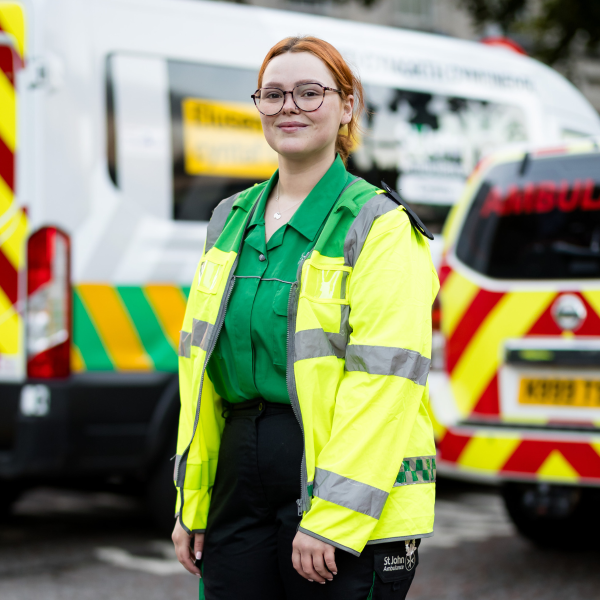 St John ambulance Cymru volunteer wearing green uniform and a high visibility yellow jacket. She has glasses and tied back red hair and is smiling.