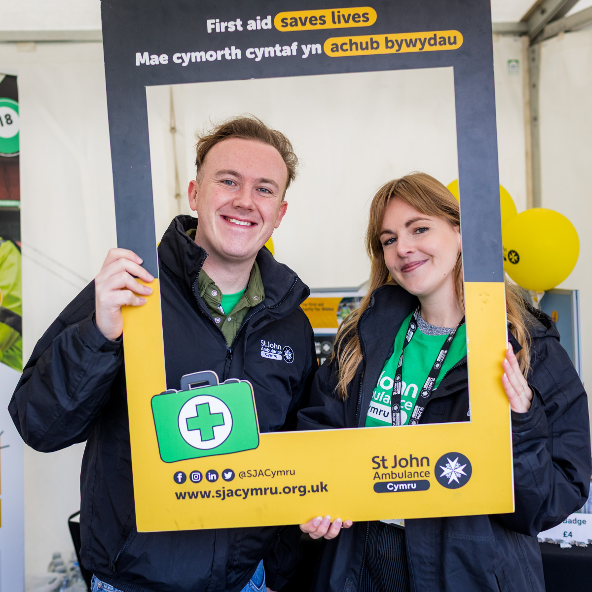 Two St John Ambulance staff Members smiling, holding up a board with the St John Ambulance logo and strapline 'first aid saves lives'.