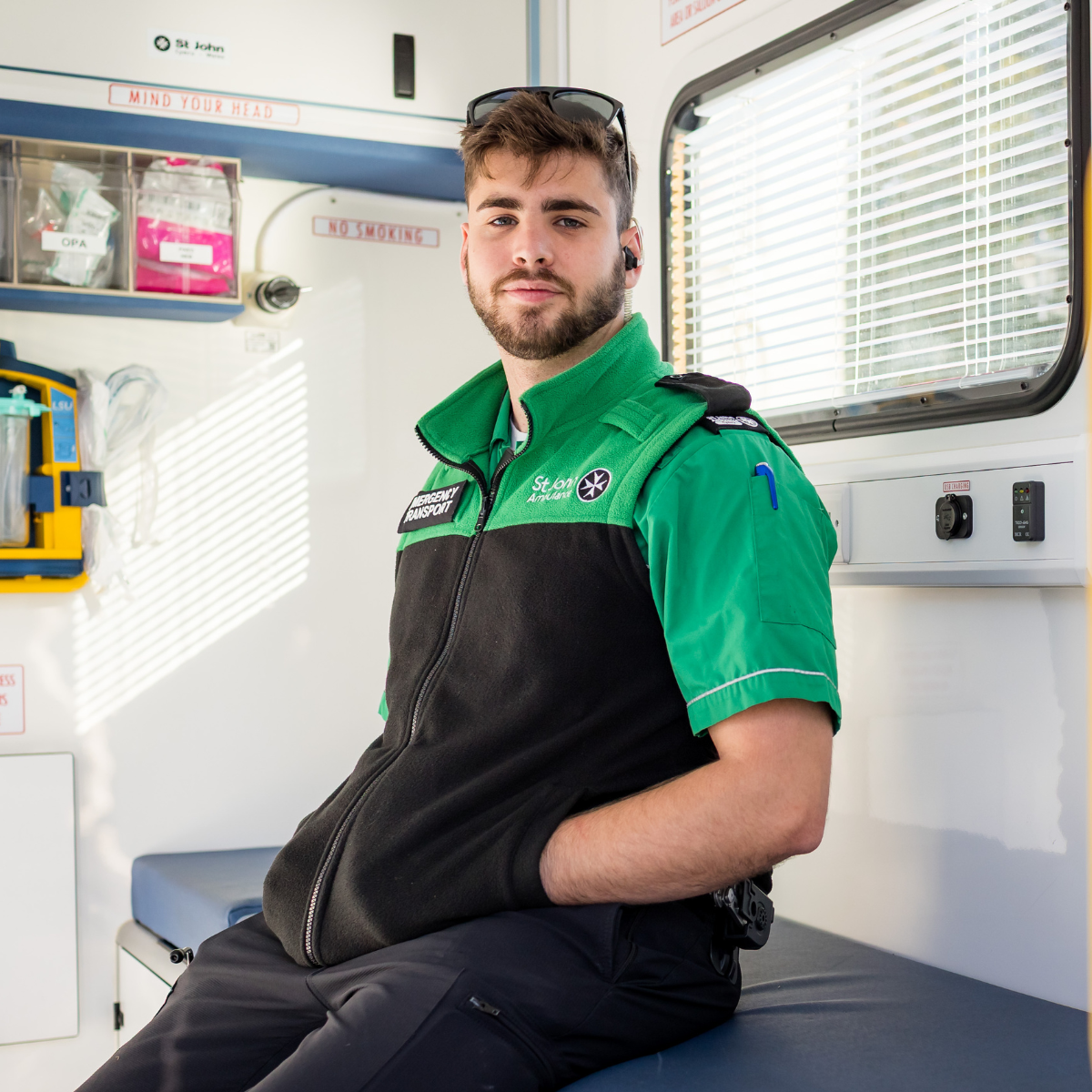 St John Ambulance Cymru staff member sitting on a medical table inside a St John Ambulance Cymru Ambulance. He is wearing green uniform and smiling at the camera. On the uniform, he has a badge stating he is Emergency Transport staff.