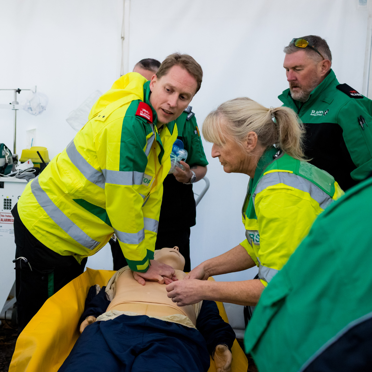St John Ambulance Cymru volunteers practising CPR first aid on a training manikin. There is four volunteers, all in uniform. The volunteer doctor has the heel of one hand in the centre of the training manikins chest. His other hand is placed on top of the first.