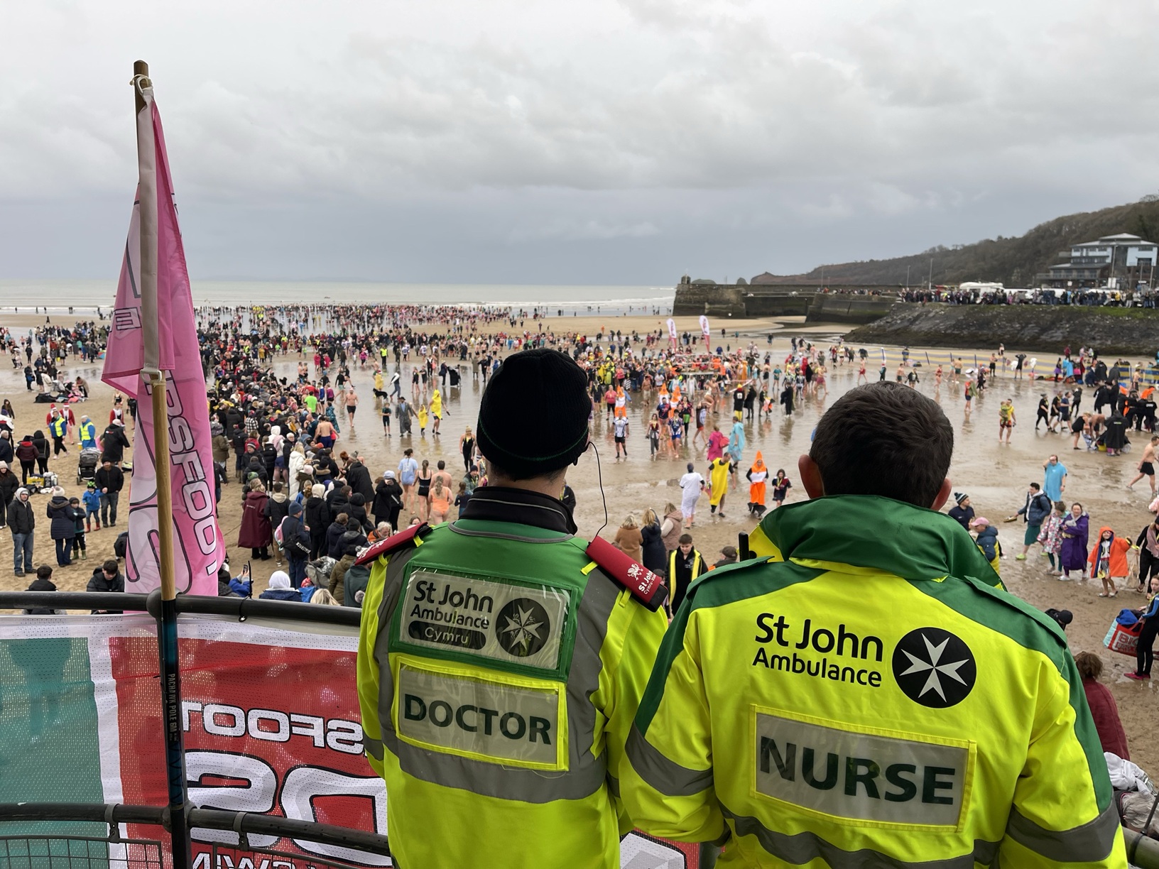 Two St John Ambulance Cymru volunteers wearing yellow and green uniforms overlooking an event taking place on a beach