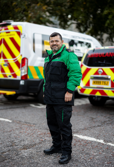 St John ambulance Cymru volunteer wearing green uniform. He is standing in car park with Ambulances behind him. He is smiling and has a beard.