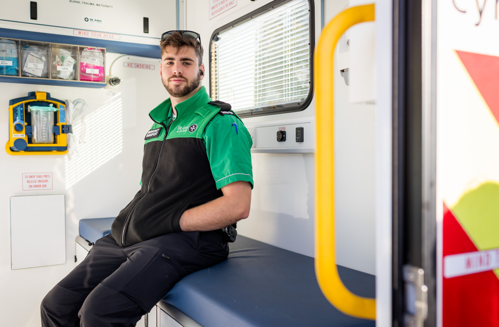 St John Ambulance Cymru staff member sitting on a medical table inside a St John Ambulance Cymru Ambulance. He is wearing green uniform and smiling at the camera. On the uniform, he has a badge stating he is Emergency Transport staff.