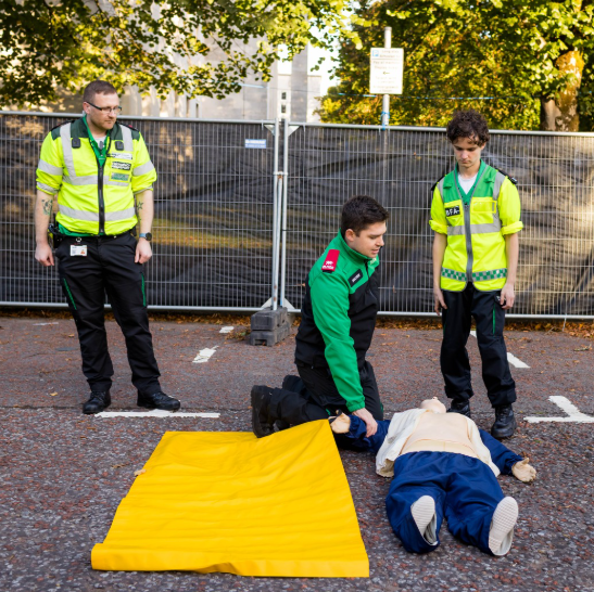 St John Ambulance Cymru volunteers practising first aid on a training manikin outdoors, with a yellow mat beside them