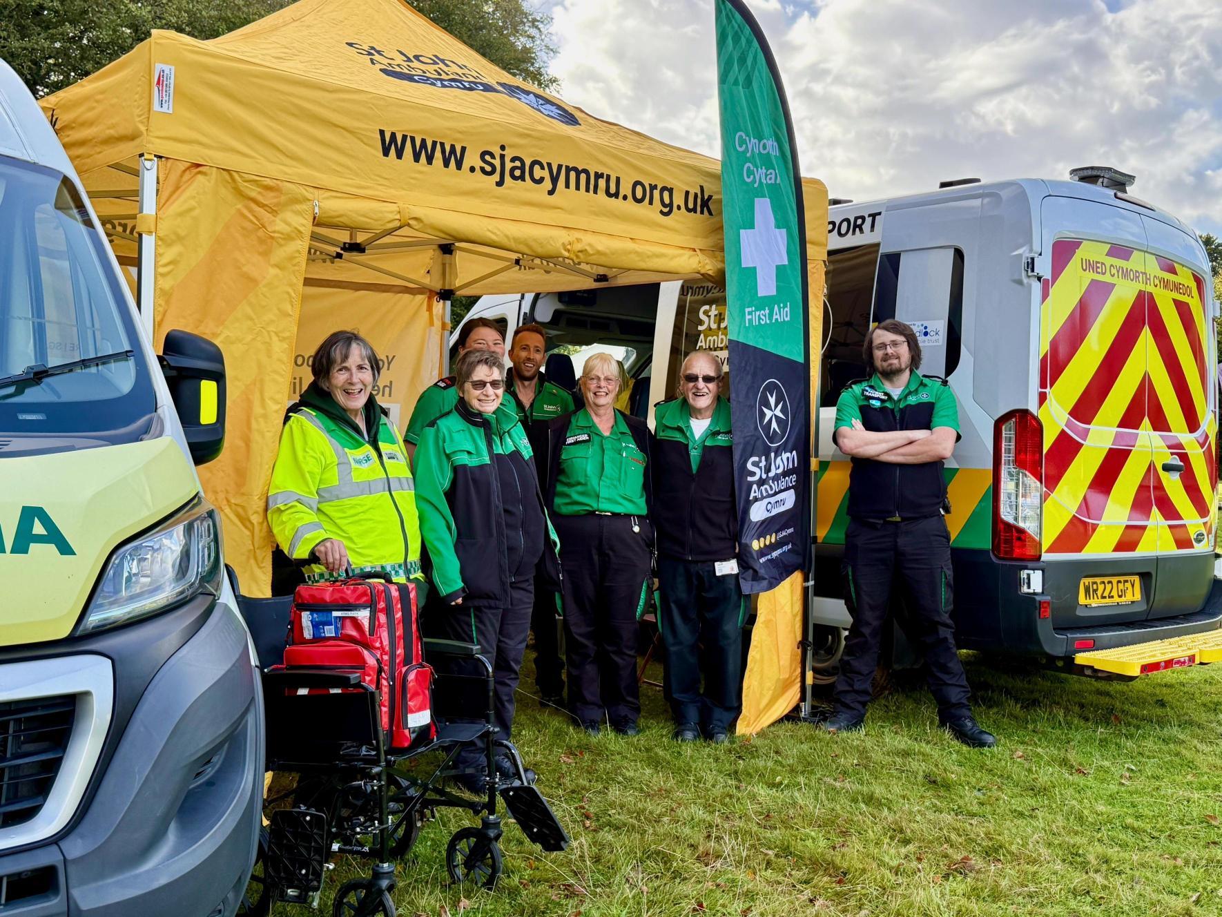 Volunteers at St Fagans Food Festival smiling, all wearing green St John Ambulance Cymru uniforms, some in high-visibility jackets, with ambulances on either side.