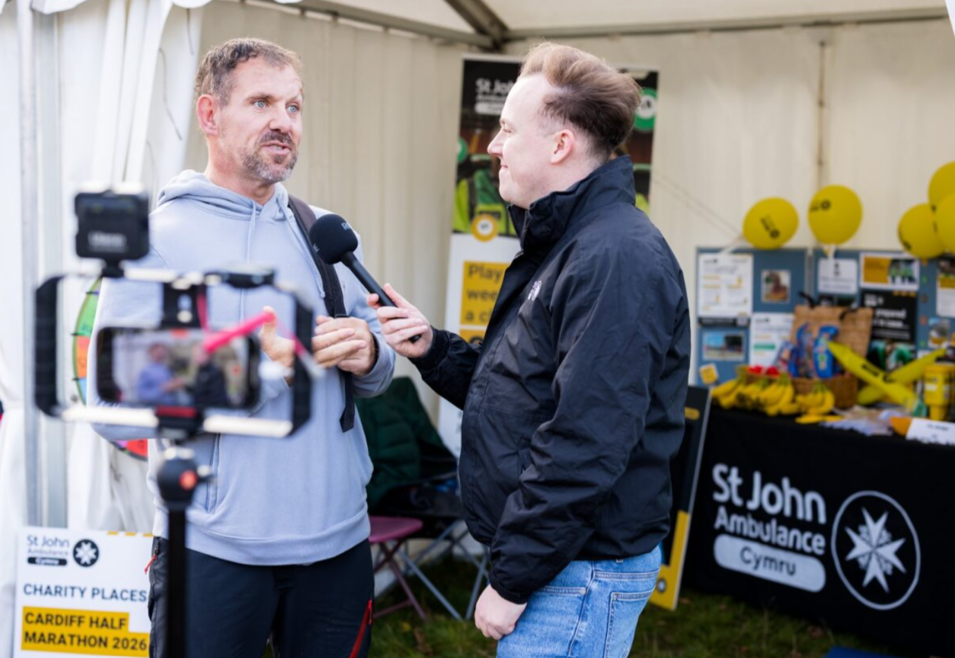 Traitors finalist and St John Ambulance Cymru ambassador Andrew Jenkins is being interviewed by a St John Ambulance Cymru staff member. They are standing in front of the St John Ambulance Cymru tent at the Cardiff Half Marathon. Behind him, in the tent are banners with the St John Ambulance Cymru logo. They are in mid conversation and Andrew is talking into a microphone.