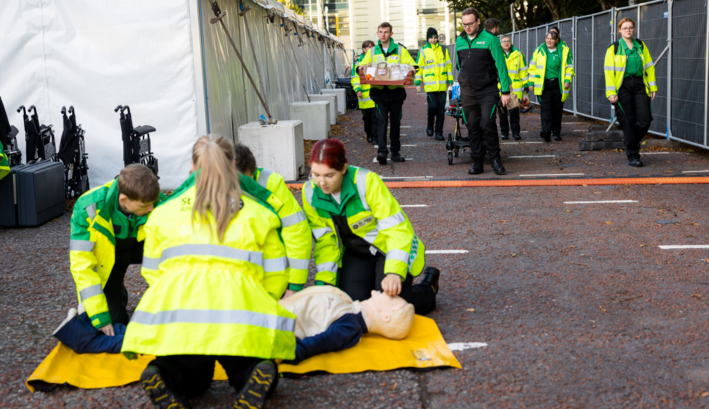Four St John Ambulance Cymru volunteers in high visibility jackets practice first aid techniques on a training manikin. They are outside and have a medical tent in the background.