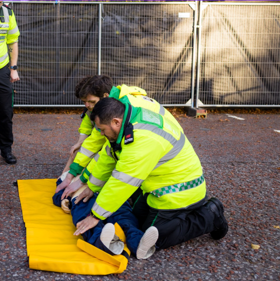 St John Ambulance Cymru volunteers practising first aid on a training manikin outdoors, on a yellow blanket.
